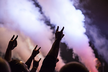 Fire arme stikker i vejret og laver håndtegn til koncert. Der er røg lyst op i lyserøde farver.